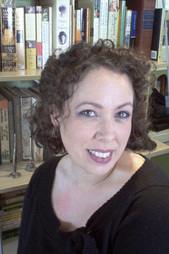 A photo of author Isobel Carr. She has dark curly hair, a pretty smile and she is wearing black. Behind her, there are books.