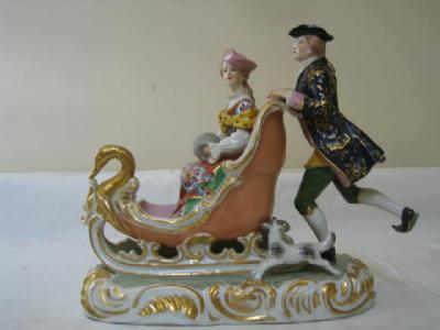 Porcelain figurine depicting and 18th century woman sitting upright in her sleigh-like sledge, while a gentleman wearing skates pushes it along the ice from behind.