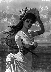 Young lady dressed in late Victorian style clothing and large rbimmed straw hat, one hand holding it against a breeze and a tennis racquet clutched under her arm.