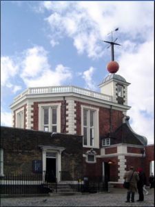 Semi-octagonal brick tower at the Greenwich Observatory with a small white wooden clock tower above at the right side, surmounted by a cross-shaped pole with a large gold ball at the base, resting at the roofline.