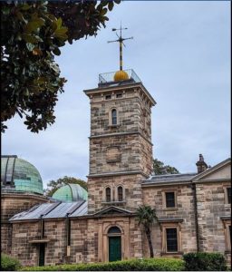 The Sydney Observatory is a large granite stone building with a central tower of four stories. Affixed to the roof of the tower is the typical cross-shaped support pole with a large golden ball resting at its base.